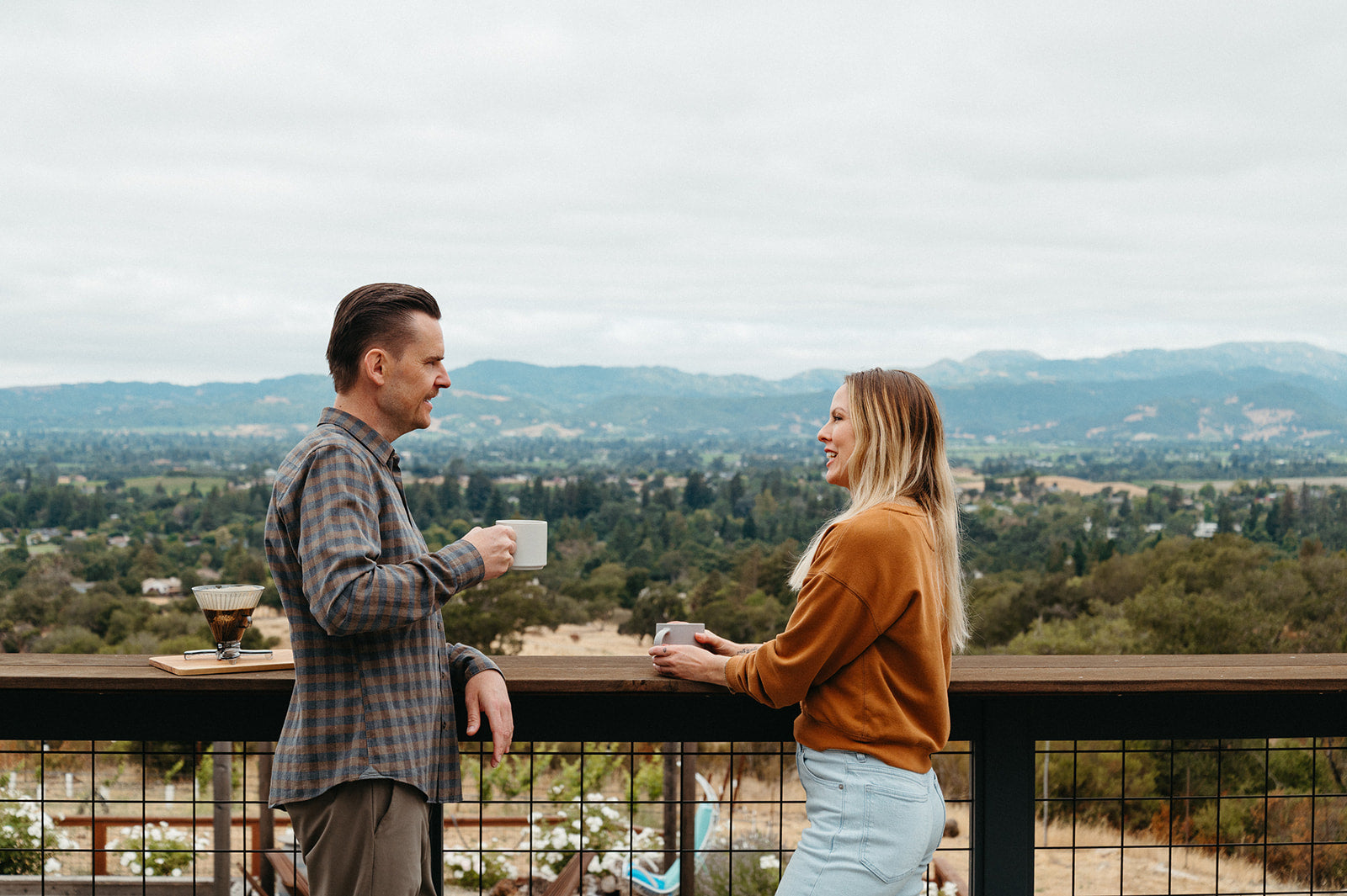 Man and woman standing on a balcony overlooking a scenic landscape with mountains.