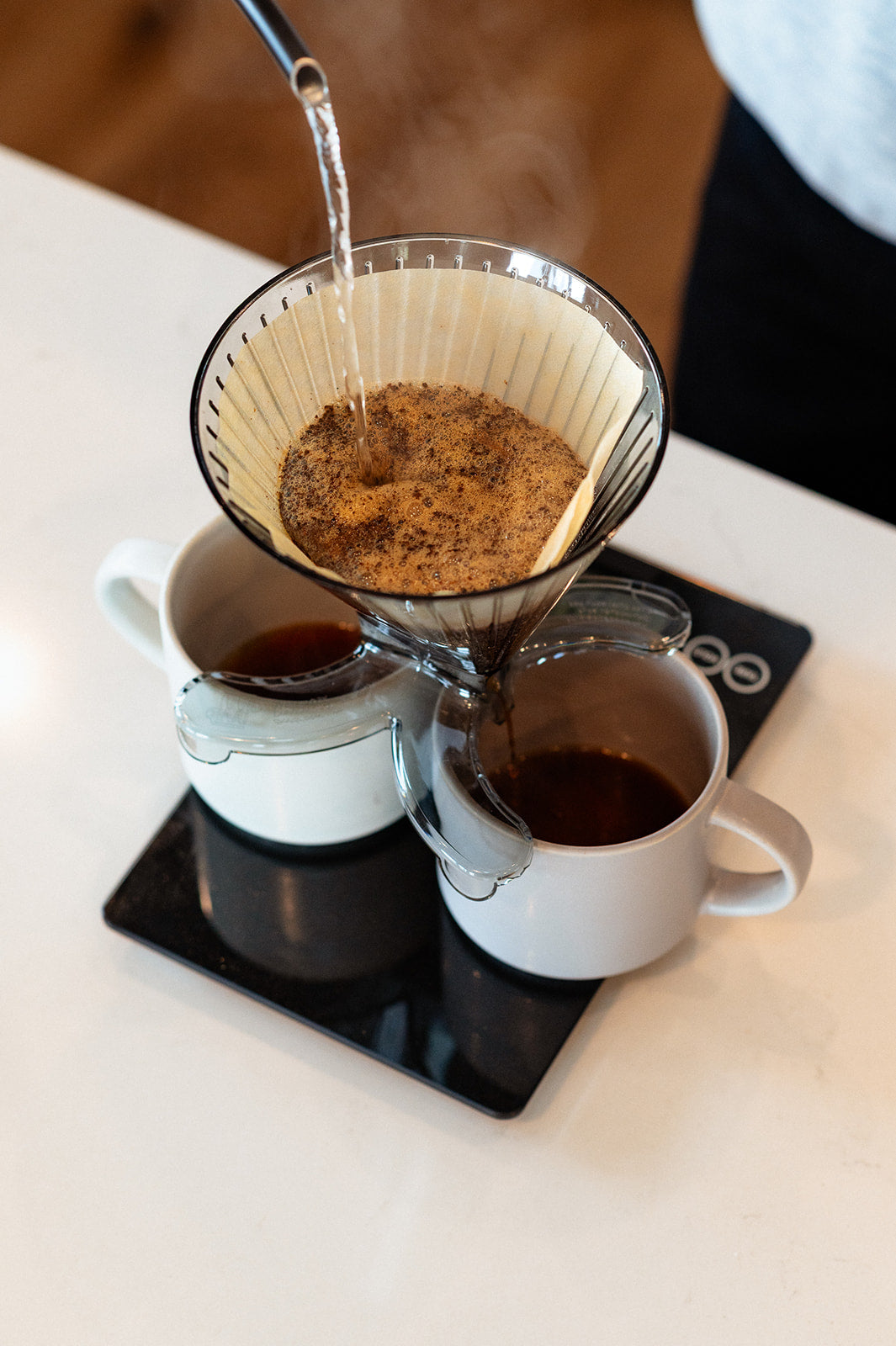 Coffee being made with a Divvi pourover flowing evenly into two mugs that are sitting atop a coffee scale on a white counter.
